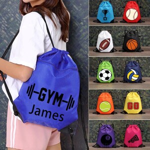 Könnte beinhalten: Ein blauer Turnbeutel mit Kordelzug und dem schwarzen Text "-GYM- James". Zusätzliche Taschen mit Sportmotiven, darunter eine Trophäe, ein Baseball, ein Football, ein Basketball, ein Fußball, ein Volleyball, ein Tennisball und eine 8-Ball.