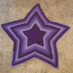 May include: A purple and lavender crocheted star-shaped blanket. The blanket is made with a variety of shades of purple yarn, creating a gradient effect.