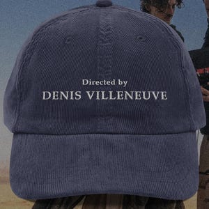 May include: A navy blue corduroy baseball cap with the text "Directed by DENIS VILLENEUVE" embroidered in white. The cap has a curved brim and a top button. The background features two people.