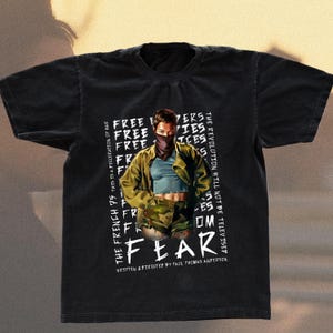 May include: Black graphic t-shirt with a central image of a person wearing a face covering and military-style clothing. The word "FEAR" is prominently displayed, along with text that reads "THE FRENCH 75 THIS IS A FEDERATION OF WAR" and "THE REVOLUTION WILL NOT BE TELEVISED".
