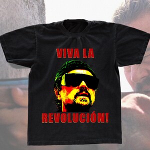 May include: Black t-shirt featuring a colorful graphic of a person wearing sunglasses and a hat. The text "VIVA LA REVOLUCIÓN!" is printed in red above and below the graphic. The shirt has a vintage, worn look.