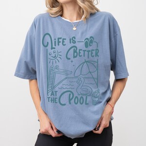 Peut inclure: T-shirt bleu avec l'inscription "Life is Better at the Pool" en vert d'eau. Le motif comprend un plongeoir, un soleil, un parasol, un flotteur cygne et une glace. Le t-shirt a des manches courtes et une coupe ample.