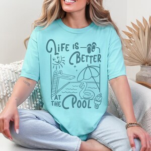 Peut inclure: T-shirt bleu clair avec l'inscription "Life is Better at the Pool". Le motif comprend un plongeoir, un soleil, un parasol, un cygne et une glace. Le t-shirt a un col rond et des manches courtes. La police est fantaisiste.