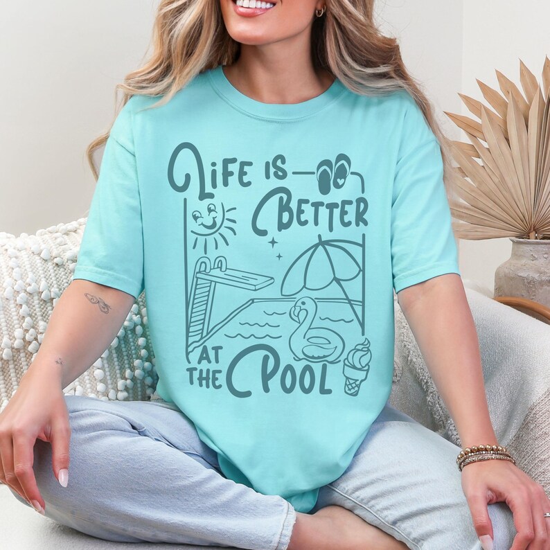 Peut inclure: T-shirt bleu clair avec l'inscription "Life is Better at the Pool". Le motif comprend un plongeoir, un soleil, un parasol, un cygne et une glace. Le t-shirt a un col rond et des manches courtes. La police est fantaisiste.