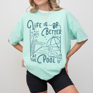 Peut inclure: T-shirt vert clair avec l'inscription "Life is Better at the Pool" en turquoise. Le motif comprend un plongeoir, un soleil, un parasol, un flotteur cygne et une glace.