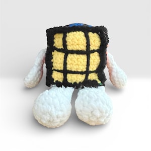 May include: A handmade crochet cube-shaped plush toy with a yellow and black grid pattern. The toy has white, fluffy arms and large, rounded feet. The top of the cube is blue. The toy is set against a plain white background.
