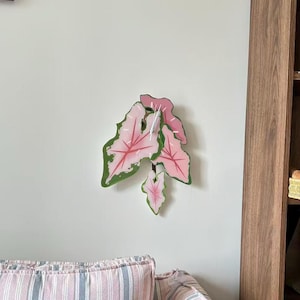 May include: A wall clock shaped like a cluster of Caladium leaves with pink centres, dark pink veins, and green borders. The clock features white hands centred on the largest leaf, displaying a botanical design with a smooth, flat surface.