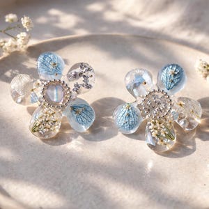 Peut inclure: Deux boucles d'oreilles en forme de fleur avec des pétales en résine transparente, chacune contenant des éléments bleus et blancs. Le centre de chaque boucle d'oreille présente un sertissage argenté avec une pierre centrale. Des fleurs blanches délicates sont disposées sur une assiette de ton neutre.
