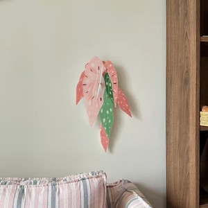 May include: A wall clock shaped like a cluster of begonia leaves in shades of pink and green with white polka dots. The clock features thin white hands and black hour markers, mounted on a plain wall above a striped sofa with a brown plush dog toy.