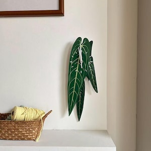 Op de afbeelding: Een wandklok in de vorm van een cluster donkergroene bladeren met opvallende lichtgroene nerven. De wijzerplaat heeft witte uurmarkeringen en dunne witte wijzers die centraal op het grootste blad zijn geplaatst voor een botanische uitstraling.