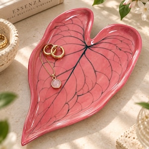 Puede incluir: Un plato decorativo de cerámica en forma de corazón de color rosa vibrante, con un patrón detallado de venas oscuras que recuerda a una hoja. El plato contiene un collar dorado con un colgante redondo y un par de pendientes de aro dorados.