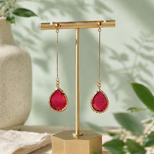 May include: A pair of long gold-tone drop earrings featuring teardrop-shaped magenta stones encased in a textured gold-tone metal frame, suspended from thin gold-tone chains attached to a gold-tone T-shaped jewelry display stand.