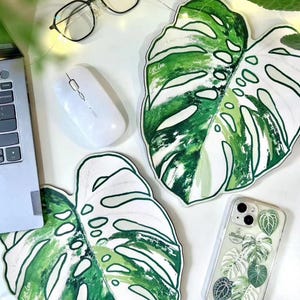 May include: Two leaf-shaped mouse pads with a green and white monstera leaf design. A white wireless mouse, eyeglasses, and a phone case with a leaf pattern are also visible.