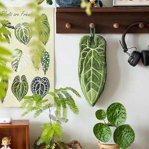 Puede incluir: Un adorno de pared verde en forma de hoja con detalles de venas blancas, suspendido de un lazo verde. Un póster botánico con varias ilustraciones de hojas cuelga cerca. Una pequeña planta en maceta se encuentra sobre un taburete de madera, y unos auriculares cuelgan de un estante de madera.