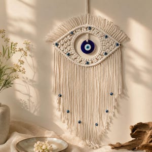 May include: A wall hanging made of off-white macrame cord featuring an eye-shaped design. The center contains a circular blue and white glass evil eye bead. Smaller blue evil eye beads are woven into the cord and attached to the long fringe.