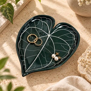 May include: A dark green ceramic trinket dish shaped like a heart-shaped leaf with white painted veins. The dish holds two gold-toned rings and a pair of white pearl stud earrings with gold-toned posts, resting on a textured surface.