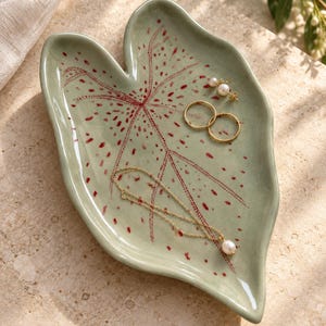 May include: A sage green ceramic trinket dish shaped like a leaf, featuring a hand-painted red dotted vein pattern. The dish holds two gold rings, a pair of small pearl stud earrings, and a delicate gold chain necklace with a single pearl pendant.