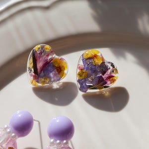 Peut inclure: Boucles d'oreilles en résine en forme de cœur avec des fleurs séchées violettes, jaunes et roses. Les boucles d'oreilles sont transparentes avec une tige dorée. D'autres boucles d'oreilles avec des perles violettes et des motifs floraux sont également visibles.