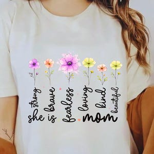Op de afbeelding: Crèmekleurig T-shirt met een bloemenontwerp en de woorden "strong, brave, fearless, loving, kind, beautiful, mom." De bloemen zijn roze, geel en oranje. De persoon draagt een bruine hoed.