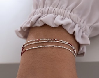 May include: Three delicate bracelets with silver beads and red accents. The bracelets are strung on a thin, dark red cord. The bracelets are stacked on top of each other.
