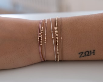 May include: A close-up of a wrist wearing multiple bracelets. The bracelets are made of thin string and have small gold beads. The string is in various colors, including pink, white, and brown. The wrist also has a tattoo that says "ZQH".