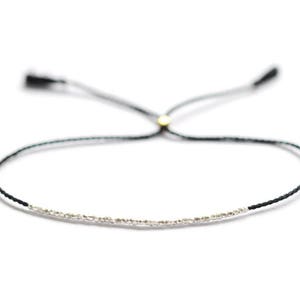 Könnte beinhalten: Ein zartes schwarzes und silbernes Armband mit einem verschiebbaren Knotenverschluss. Das Armband besteht aus einem dünnen schwarzen Band mit einer Reihe winziger silberner Perlen.
