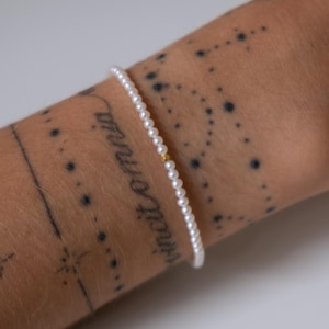 May include: A fine pearl bracelet with a small gold bead. The bracelet is strung on a thin cord. The wrist it is on has tattoos. The bracelet is a classic piece of jewellery.