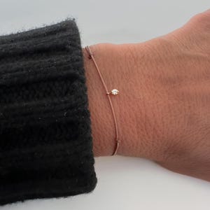 May include: A fine bracelet with a small diamond pendant. The bracelet is made of a thin, light brown cord. The diamond is set in a gold-coloured setting. The bracelet is worn on a wrist, with a black sweater sleeve in the background.