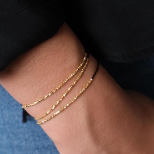 May include: A layered bracelet set with three strands of gold-coloured beads on black cords. The bracelet is worn on a wrist, with a black tassel visible. The gold beads have a faceted appearance, catching the light.