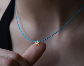 May include: A delicate gold starfish charm hangs from a turquoise blue cord necklace. The necklace is worn by a person, but the focus is on the necklace itself.