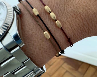 May include: Three beaded bracelets with gold-colored oval beads are displayed on a wrist, alongside a silver wristwatch. The bracelets feature dark brown, black, and burgundy cords. The beads have a textured, matte finish.
