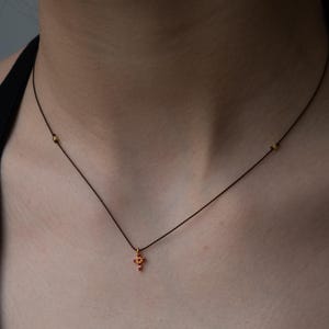 Op de afbeelding: Een delicate gouden ketting met een kruis hanger met rode edelstenen, op een zwart koord met gouden kralen.