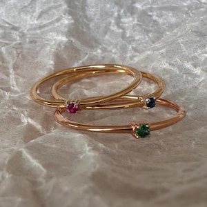 May include: Three thin gold rings, each featuring a single small round gemstone. One ring has a pink stone, one has a dark blue stone, and one has a green stone. The rings are stacked together, showcasing their delicate bands and prong settings.