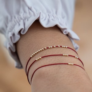 Puede incluir: Tres pulseras de cuerda roja con cuentas doradas. Las pulseras están superpuestas en la muñeca de una persona.