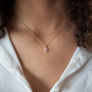 May include: A delicate gold necklace with a small hamsa hand pendant adorned with sparkling white stones and a pink gemstone in the center.