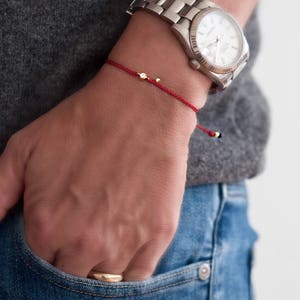 May include: A red string bracelet with small gold beads. The bracelet is worn on a person's wrist.