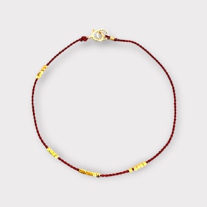 May include: A minimalist bracelet featuring a deep red cord and small, gold-colored square beads. The bracelet has a gold-colored clasp. The design is simple and elegant, suitable for everyday wear.