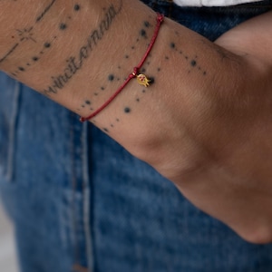 May include: A red string bracelet featuring a small gold-coloured pomegranate charm. The bracelet is worn on a wrist with visible tattoos, set against a backdrop of blue denim.