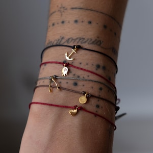 Può includere: Una collezione di braccialetti con ciondoli dorati con cordini neri, rossi e grigi. I ciondoli includono un'ancora, un melograno, un ferro di cavallo, un cerchio e un cuore. I braccialetti sono annodati con nodi regolabili.