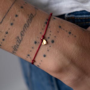 Puede incluir: Una pulsera de hilo rojo con un pequeño dije de corazón dorado. La pulsera está atada alrededor de una muñeca con un tatuaje que dice "vincit omnia". La piel tiene pequeñas pecas oscuras.