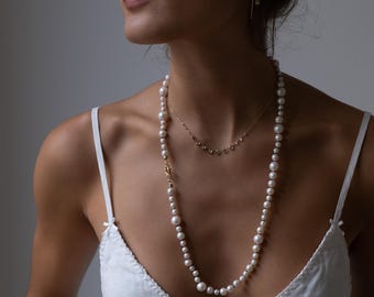 May include: A long, white pearl necklace with a gold clasp and a shorter, delicate gold necklace with small, round accents. The necklaces are worn over a white camisole.