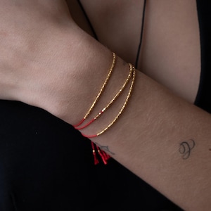 May include: Three gold beaded bracelets with red string ties. The bracelets are stacked on a person's wrist.