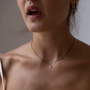 以下が含まれることがあります： 小さなゴールドのペンダントと2つのゴールドビーズが付いた黒いコードの繊細なネックレス。ネックレスは首の周りに着用されています。画像はネックレスと上半身のクローズアップを示しています。