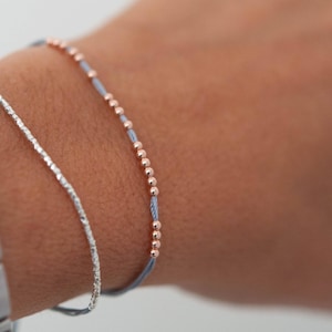May include: Two bracelets on a wrist. One bracelet is a thin silver chain, the other is a blue cord with small rose gold beads.