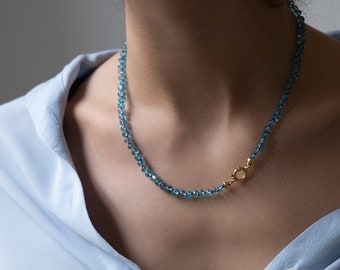 May include: A blue gemstone necklace with a gold clasp. The necklace is made of small, round beads and is worn by a person.