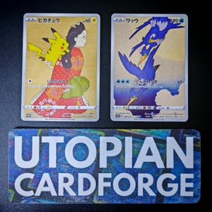 May include: Two Japanese Pokémon trading cards featuring artistic illustrations. The left card shows Pikachu with a person in a red floral kimono. The right card depicts blue birds in flight. Below them is a rectangular sign reading UTOPIAN CARDFORGE.