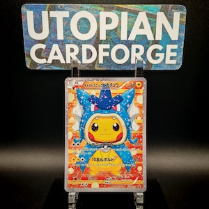 May include: A Pokémon trading card featuring Pikachu in a blue and white costume. The card has Japanese text and a yellow and orange background. The card is held in a clear plastic holder, with a sign above that reads "UTOPIAN CARDFORGE."