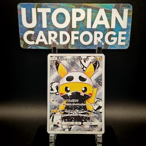 May include: A Pokemon trading card featuring a yellow Pikachu character wearing a black and white skull-themed mask and hat. The card has Japanese text and is displayed on a clear stand, with a sign above that reads "UTOPIAN CARDFORGE."