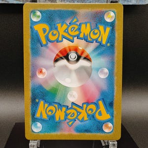 May include: A Pok&eacute;mon trading card featuring a vibrant design. The card's background is a gradient of blue and purple, with a central Pok&eacute; Ball design. The word "Pok&eacute;mon" is in yellow, and the card has a gold border.