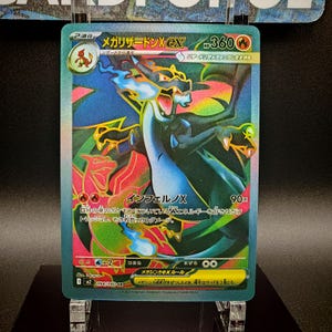 May include: A holographic Charizard EX trading card. The card features a detailed illustration of a dragon-like creature with blue flames and a red and gold outline. Japanese text is visible on the card.